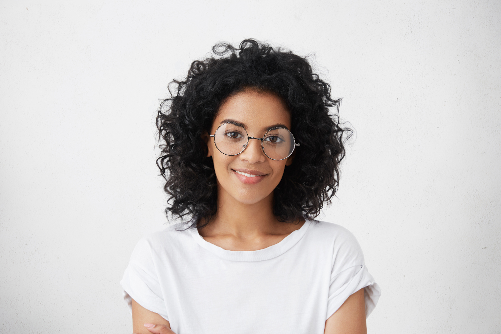 Close up studio shot of beautiful young mixed race woman model with curly dark hair looking at camera with charming cute smile while posing against white blank copy space wall for your content