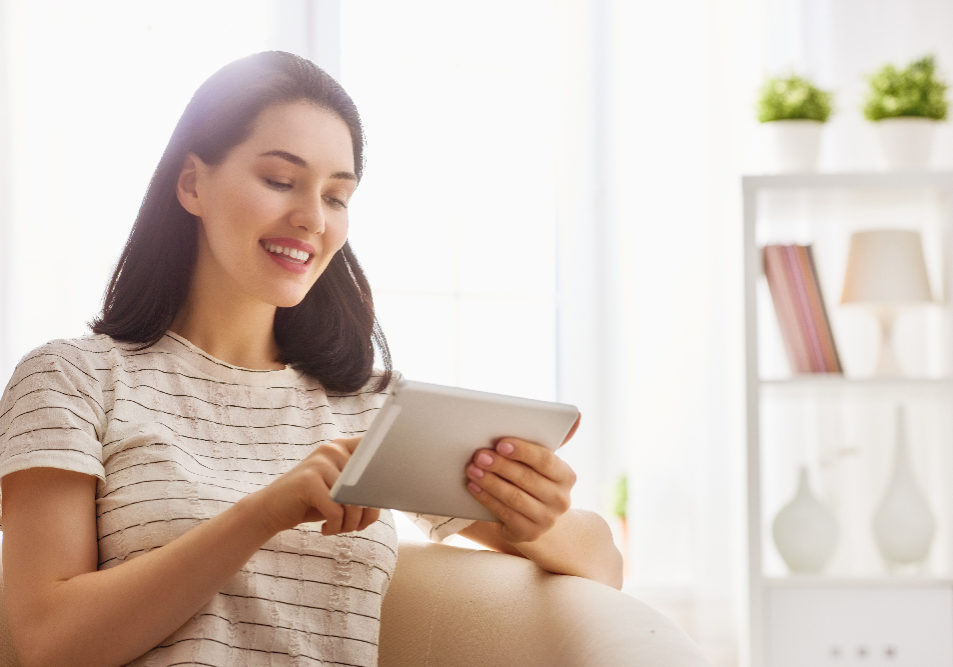 Happy beautiful woman watching videos or enjoying entertainment content on a tablet sitting on the sofa in the house.